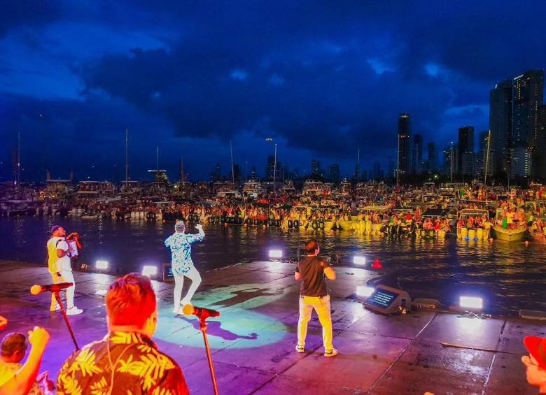Espectáculo en un muelle de noche con músicos tocando y un público sentado en barcos y plataformas cerca del agua.