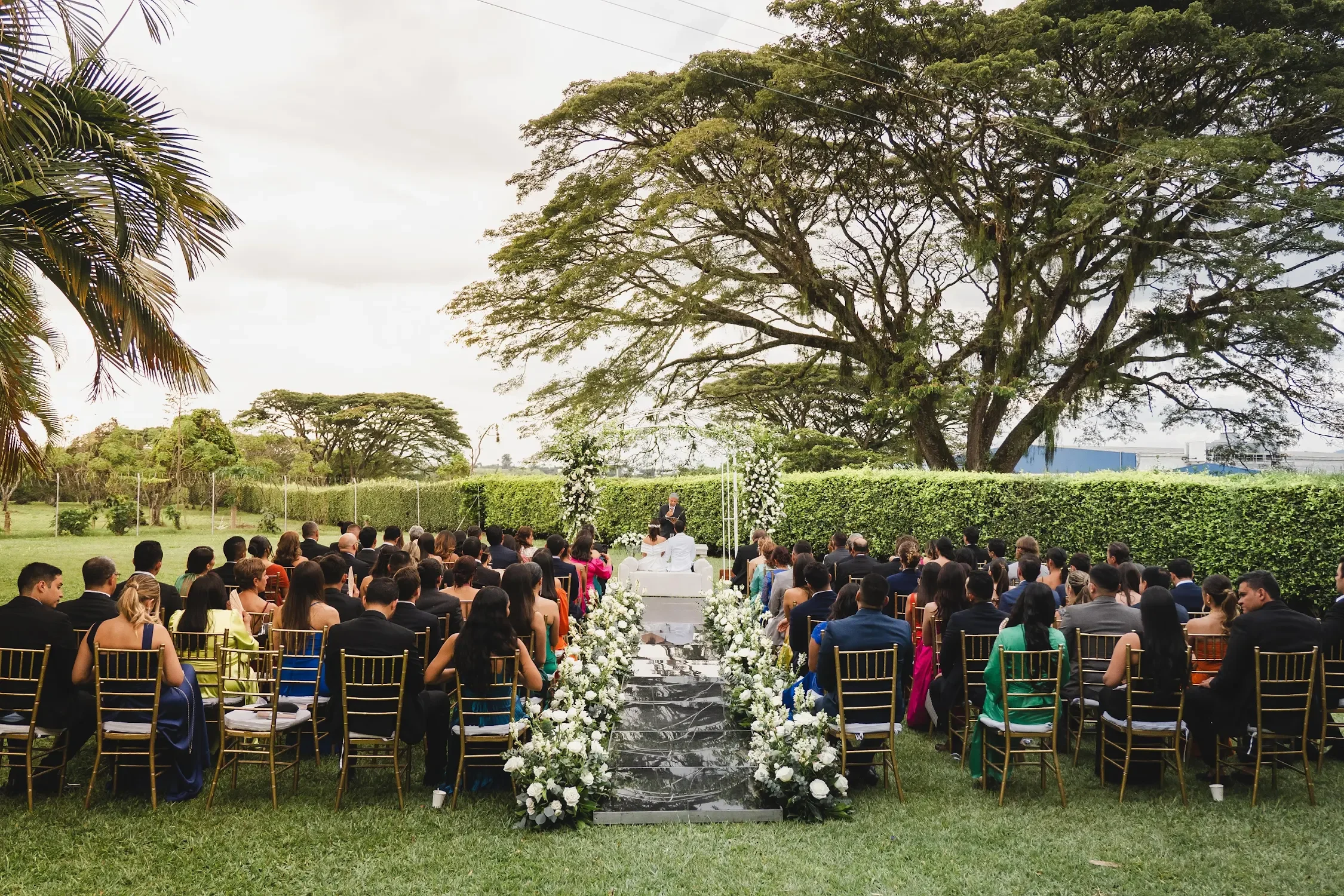 Una ceremonia de boda al aire libre con muchas personas sentadas mirando a la pareja en el altar, rodeada de árboles y un fondo de cielo nublado.