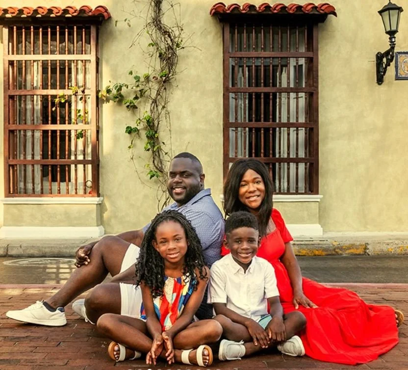 Una familia de cuatro personas sentada en el suelo frente a una pared de color beige con ventanas de barro y una lámpara de calle en la esquina derecha. La madre lleva un vestido rojo y los hijos están vestidos informalmente, todos sonrientes.