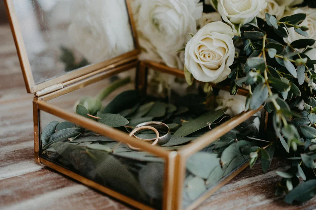 Caja de cristal con anillos dentro, rodeada de hojas verdes y flores blancas.
