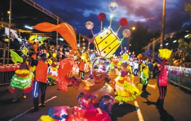 Una mujer bailando en una fiesta con vestimenta colorida y un enorme sombrero decorado con luces y globos, rodeada de otras personas vestidas con trajes llamativos, en una calle con cielo al atardecer y luces de la ciudad.