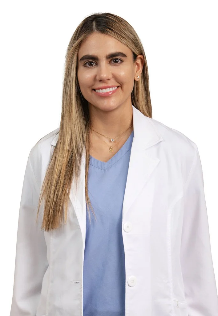 Mujer con cabello rubio, sonriendo, con bata blanca y uniforme azul, en posición frontal contra fondo blanco.