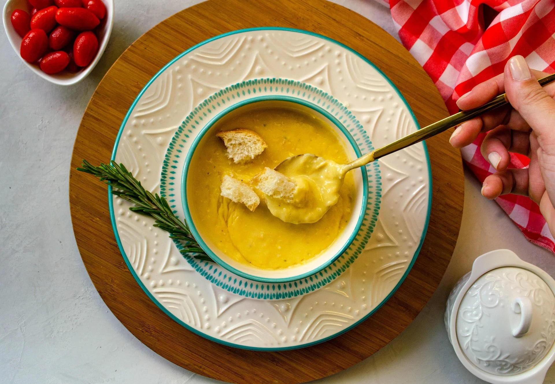 Sopa de papa con trozos de pan y un ramito de romero decorativo, en un plato con diseño. A la izquierda, un bol con jitomates cherry. A la derecha, una mano con una cuchara dorada, y una linda tela a cuadros rojos y blancos.