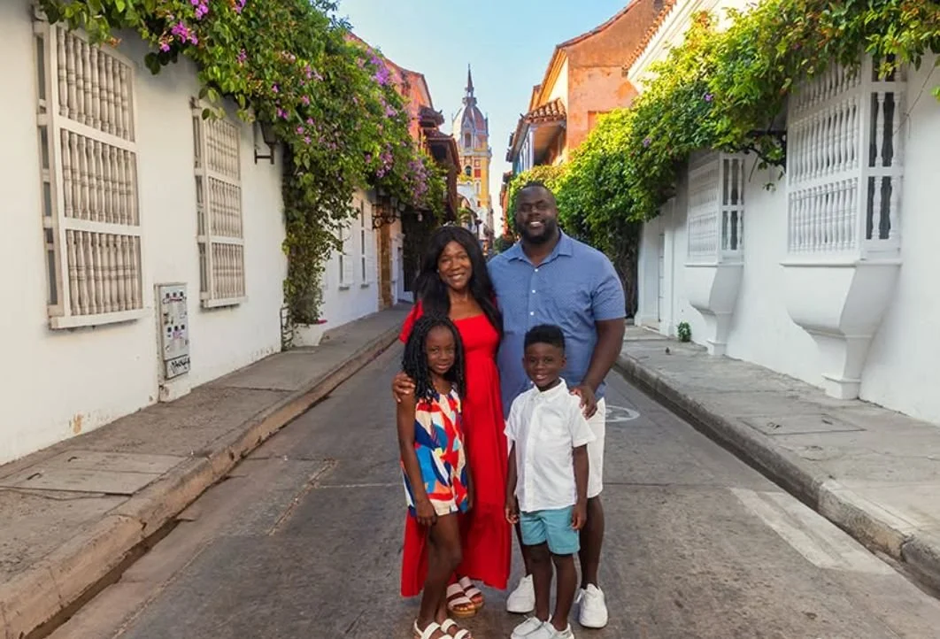 Familia de cuatro personas en una calle estrecha con casas blancas y vegetación, al fondo una iglesia con torre