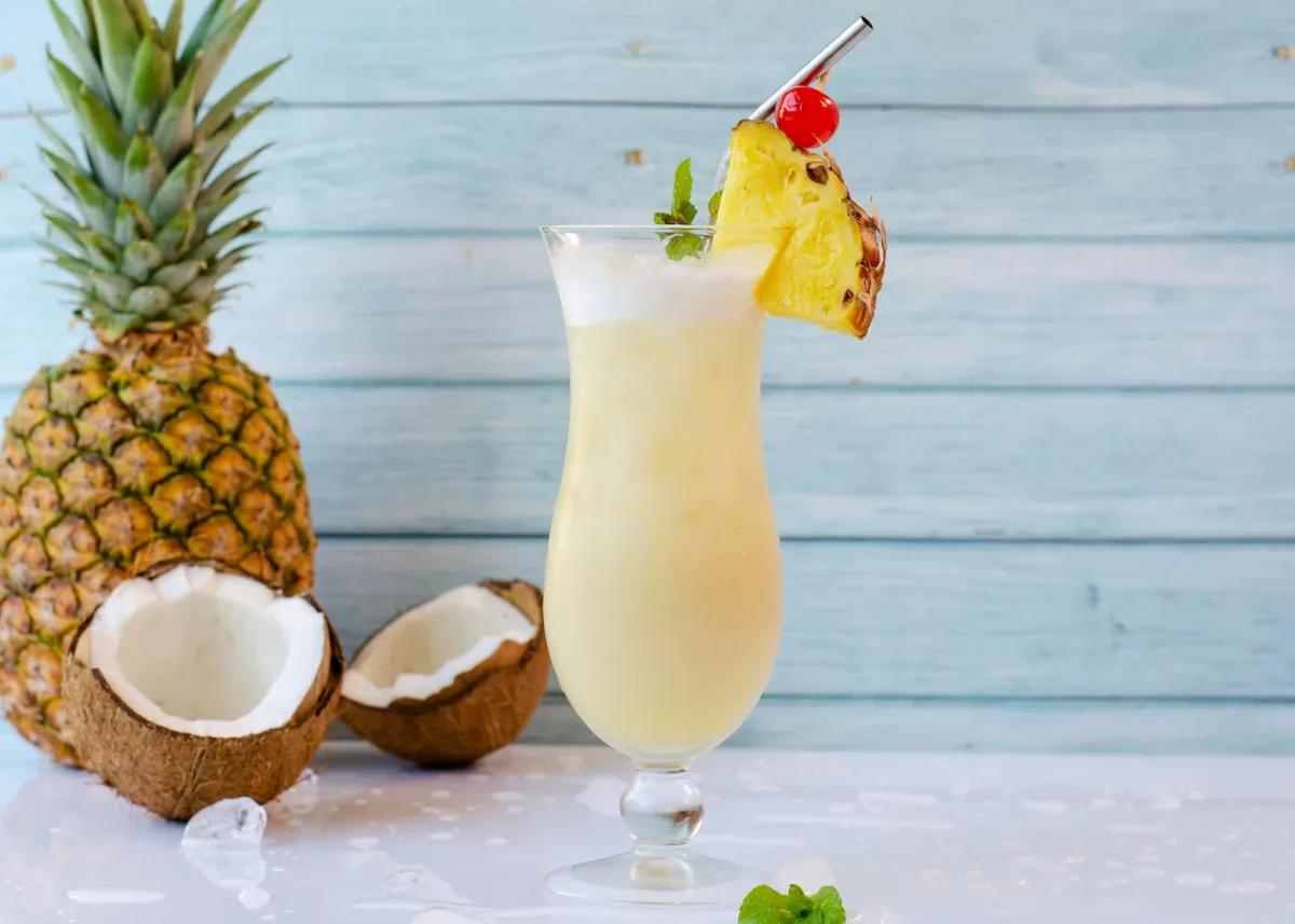 Cóctel tropical con piña, cereza y menta adornando, junto a una piña y cocolos en la mesa con fondo de tablas de madera azul.