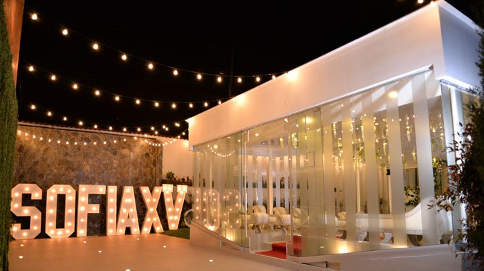 Decoración moderna y elegante para un evento nocturno, con letras grandes de luces que dicen 'SOFIAXY', cortinas de cristal y cadenas de luces en el cielo.
