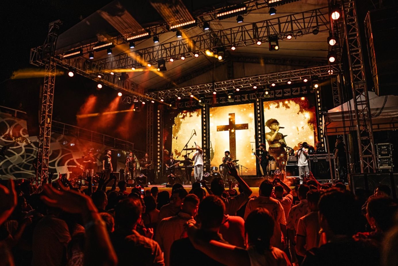Concierto en un escenario con una cruz grande y una pantalla que muestra a una persona cantando, público frente al escenario.