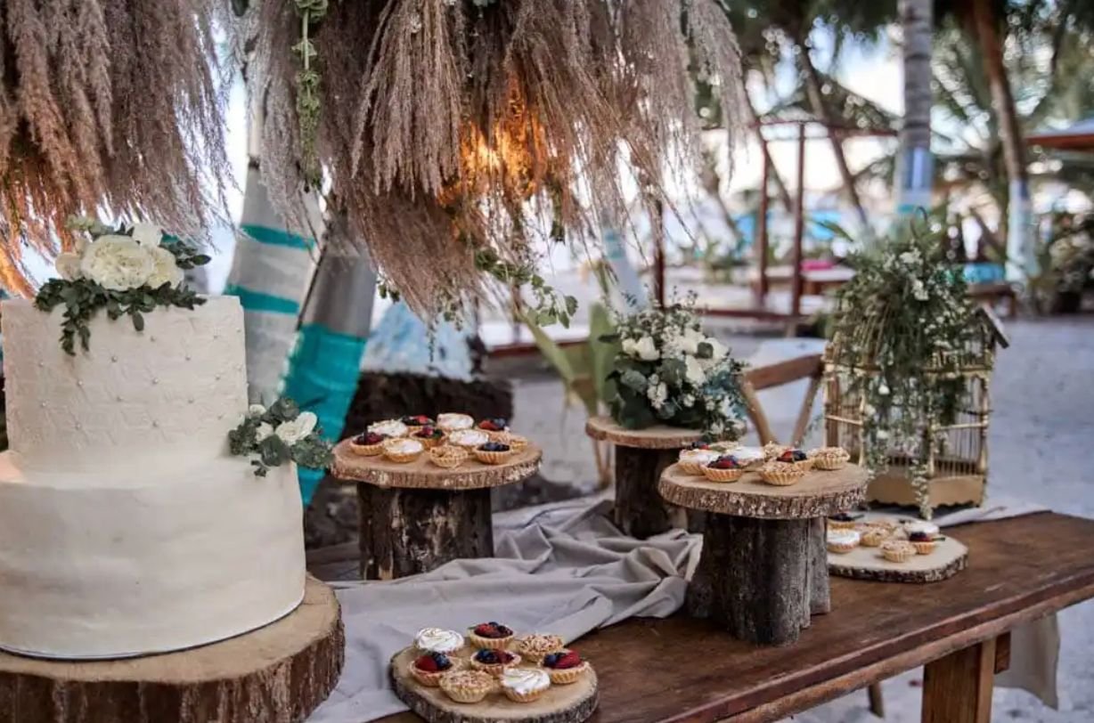 Pastel de boda blanco decorado con flores, rodeado de pequeños aperitivos en bandejas de madera, en un entorno al aire libre con árboles y decoraciones rústicas.