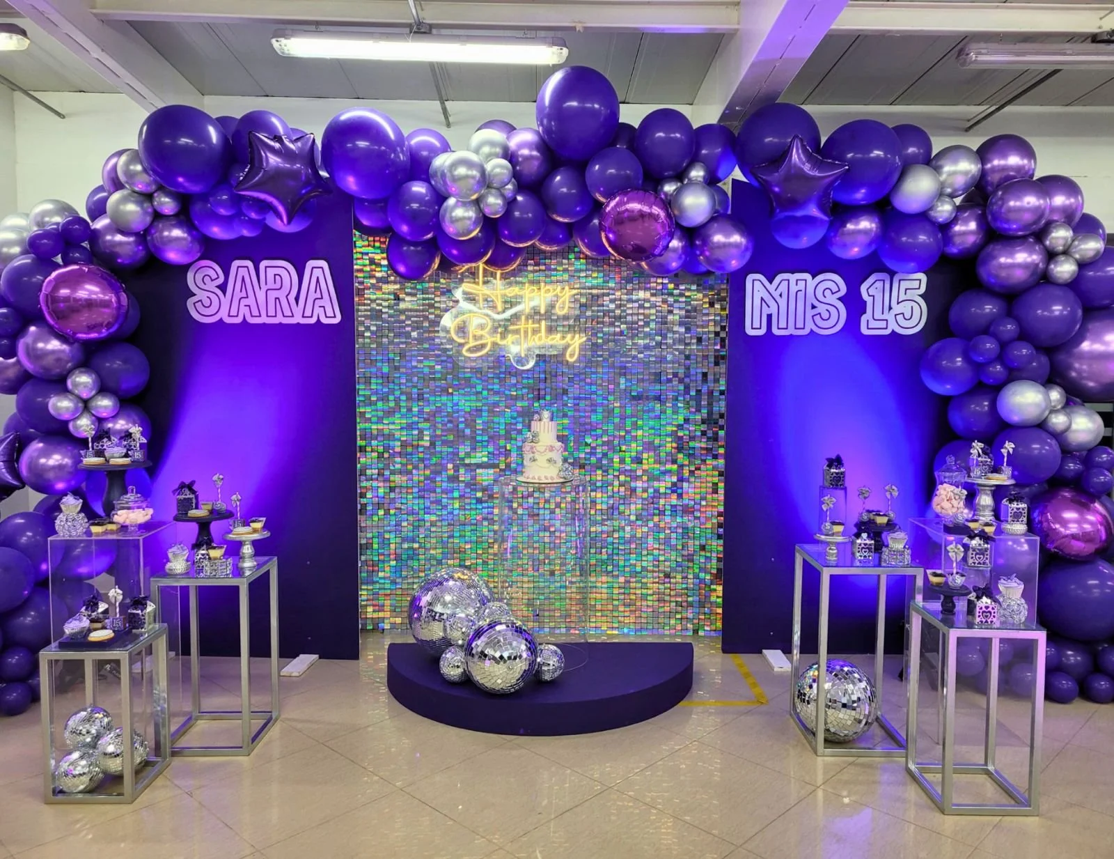 Decoración de fiesta de cumpleaños con globos morados y plateados, un pastel con velas, y un fondo brillante con la frase 'Happy Birthday'. Nombres 'Sara' y 'Mis 15' en letras grandes, varias mesas con dulces y adornos están en los lados.