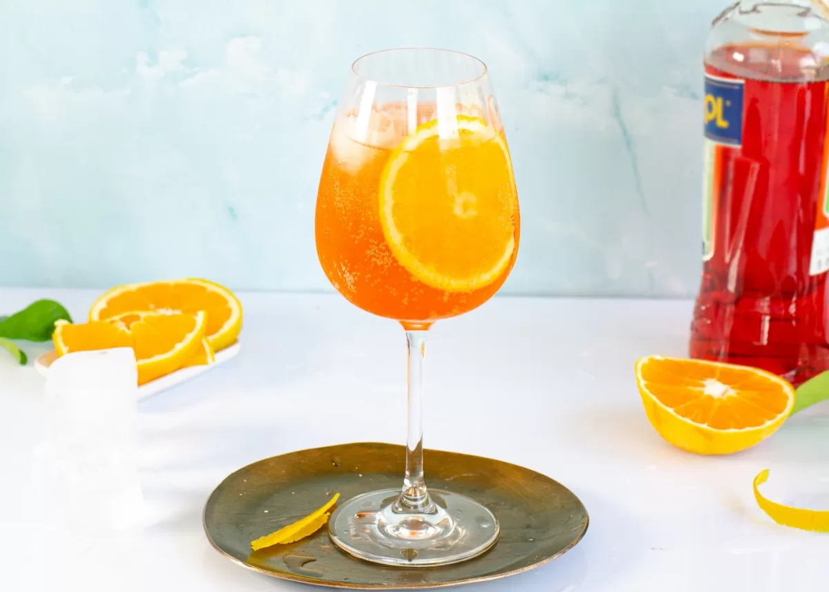 Vaso de cóctel con bebida anaranjada y rodaja de naranja, rodeado de rodajas de naranja y una botella de jarabe de color rojo al fondo.