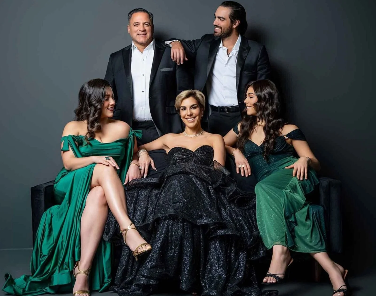 Grupo de seis personas vestidas elegantemente, cinco mujeres con vestidos largos y dos hombres con trajes, posando en un fondo oscuro.