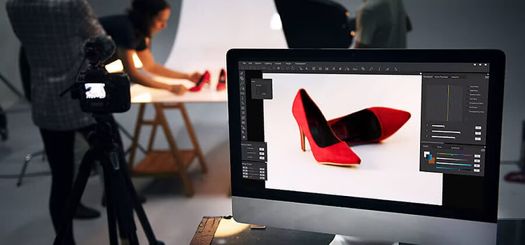 Un monitor de computadora mostrando un par de zapatos rojos con tacón en un programa de diseño, en un estudio con varias personas trabajando.
