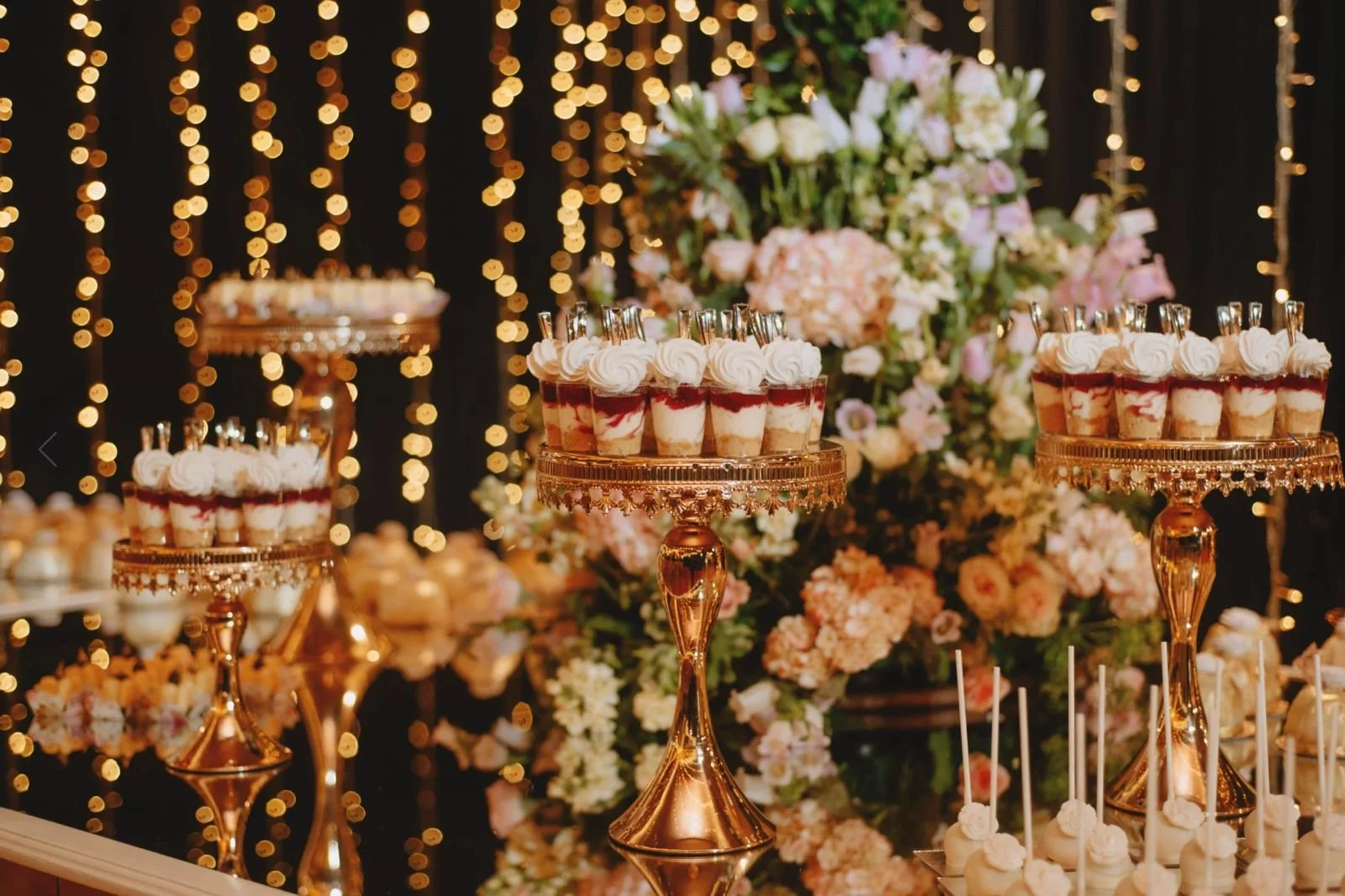 Mesas decoradas con pequeños postres en copas de cristal y flores en el fondo, en un evento elegante con luces doradas.