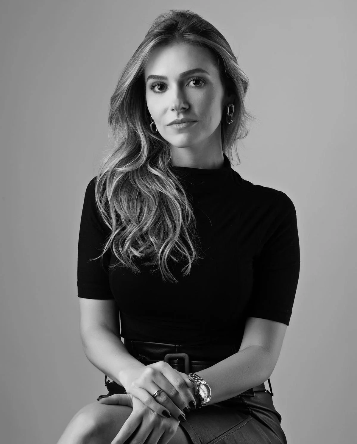 Retrato en blanco y negro de una mujer con cabello largo y rizado, con una expresión serena, vestida con un top de manga corta y accesorios, sentada contra fondo gris.
