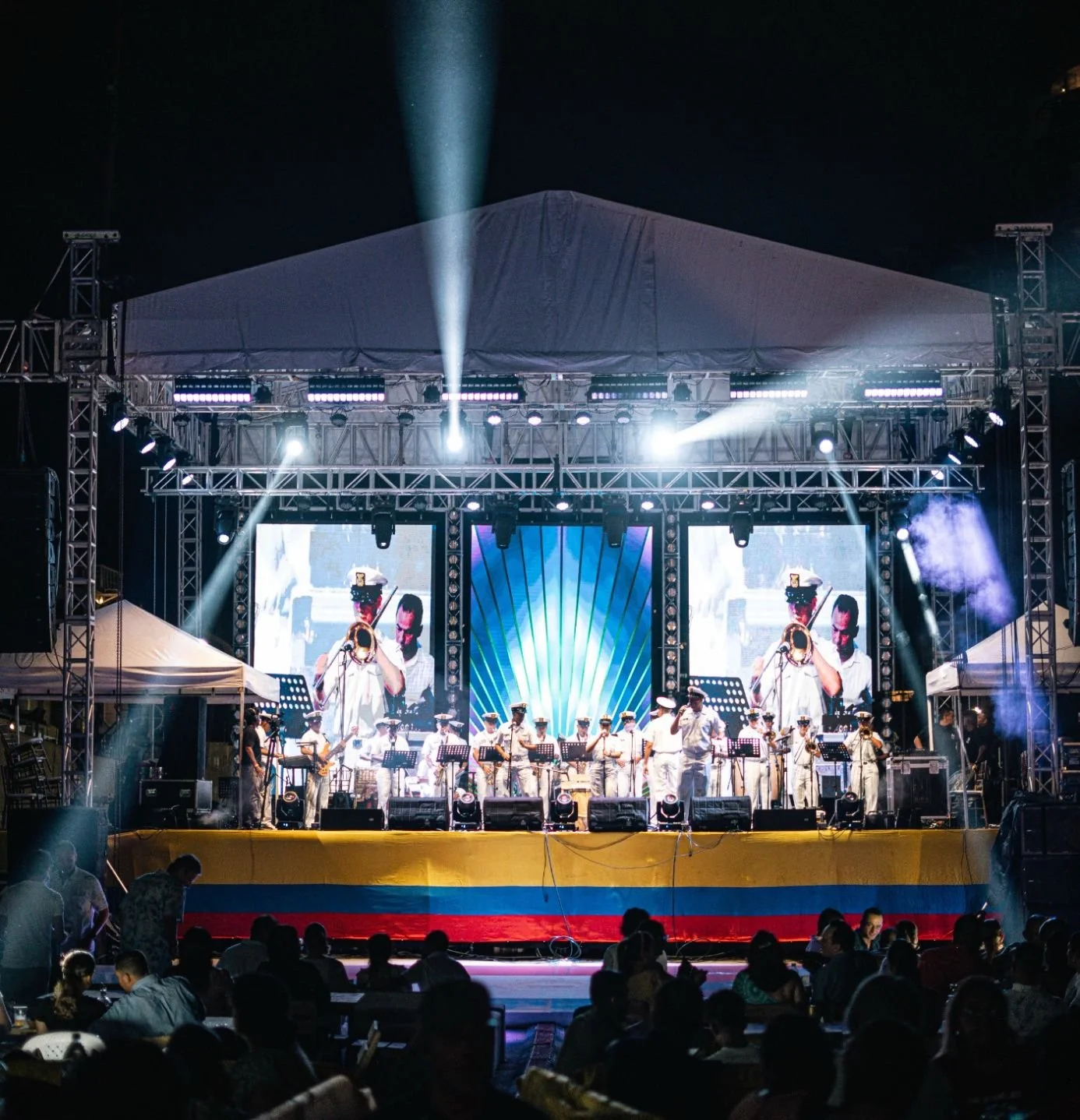 Concierto musical en una tarima al aire libre, con músicos tocando instrumentos y espectadores en primer plano, iluminación teatral y pantallas grandes funcionando atrás, con la bandera de Colombia en la parte frontal del escenario.