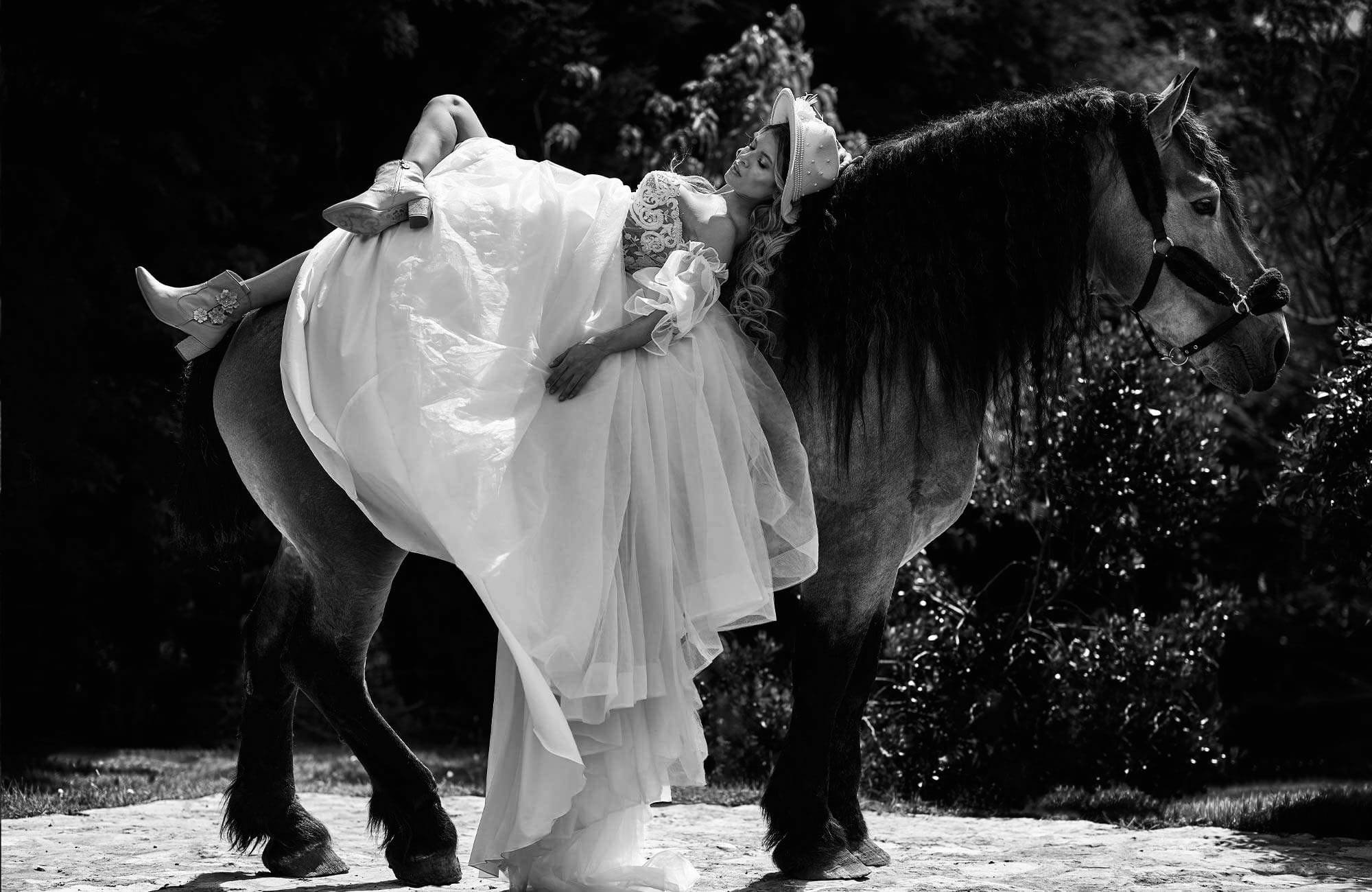 Mujer con vestido largo y sombrero descansando en un caballo en un entorno natural, en blanco y negro.