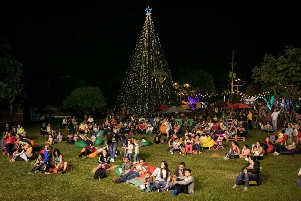 Personas reunidas en un parque de noche con un gran árbol de Navidad decorado con luces y una estrella en la cima, rodeado de juguetes y decoración navideña.