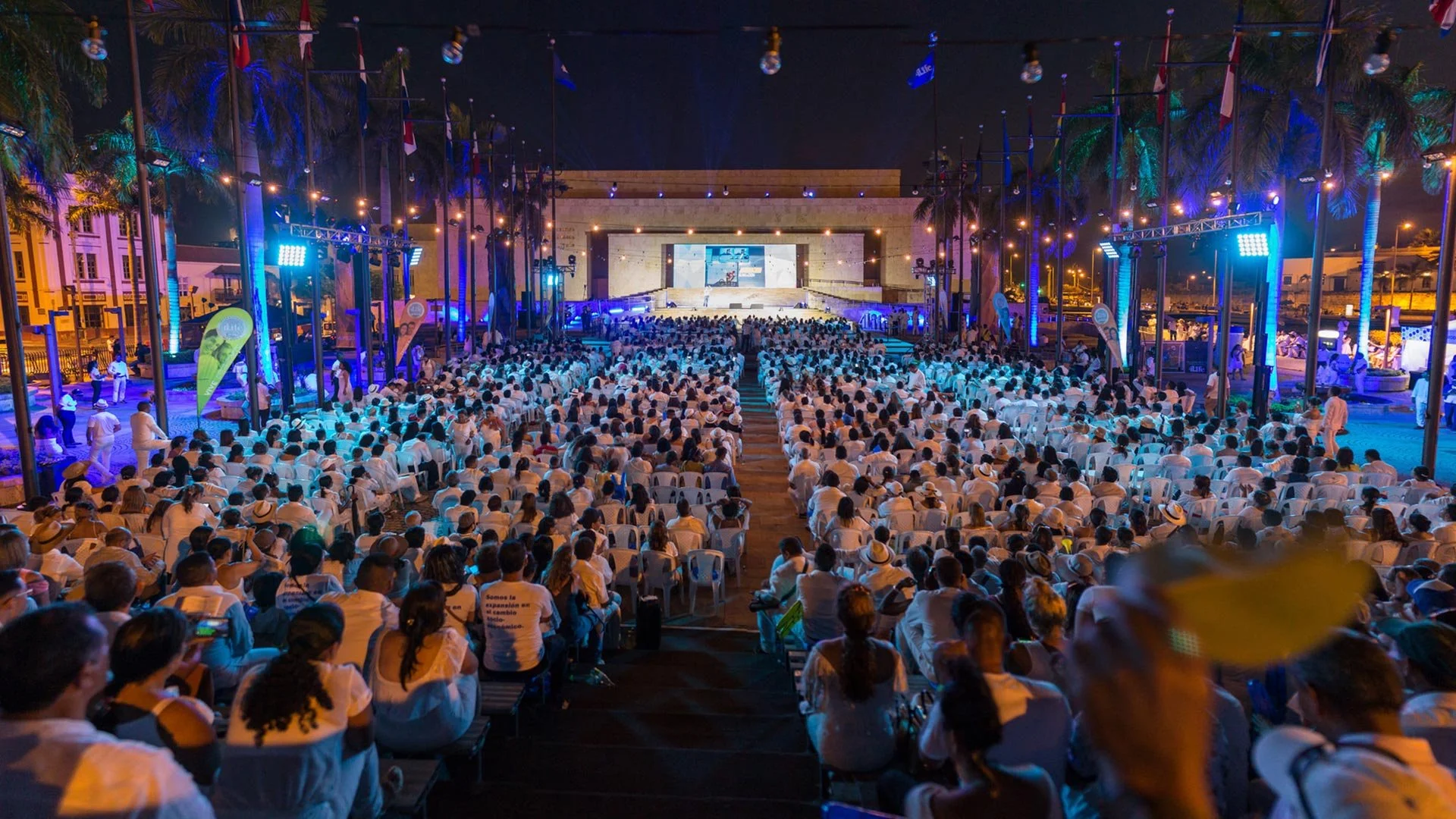 Una gran multitud de personas vestidas de blanco asisten a un evento nocturno al aire libre con escenario y pantallas, rodeado de palmeras y banderas.