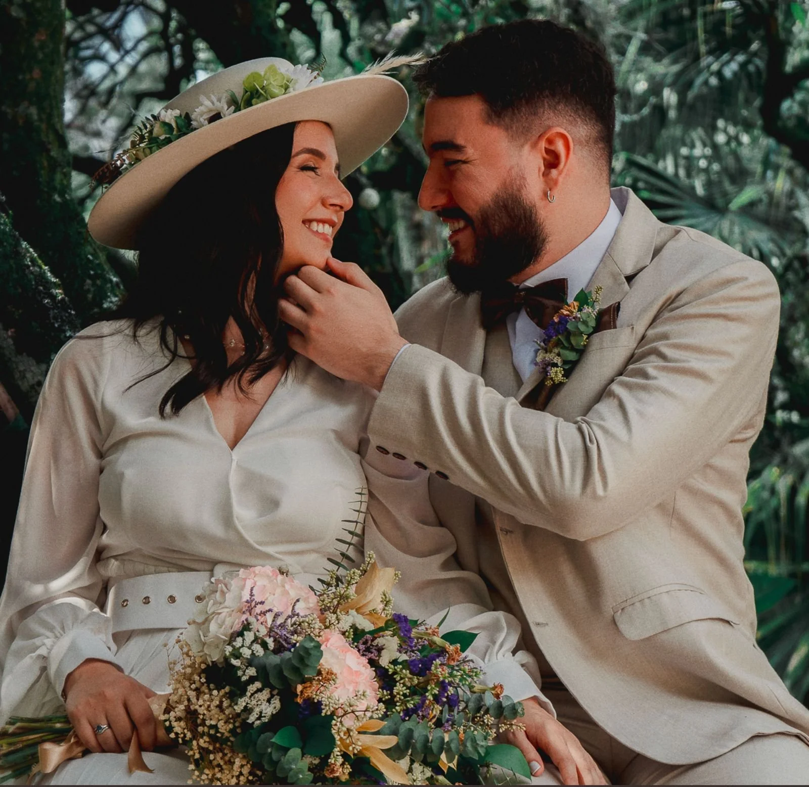 Pareja de esposos en una boda, la mujer lleva un vestido blanco y un sombrero con flores, y sostiene un ramo de flores. El hombre viste un traje beige y tiene barba, mirando con cariño a la mujer.