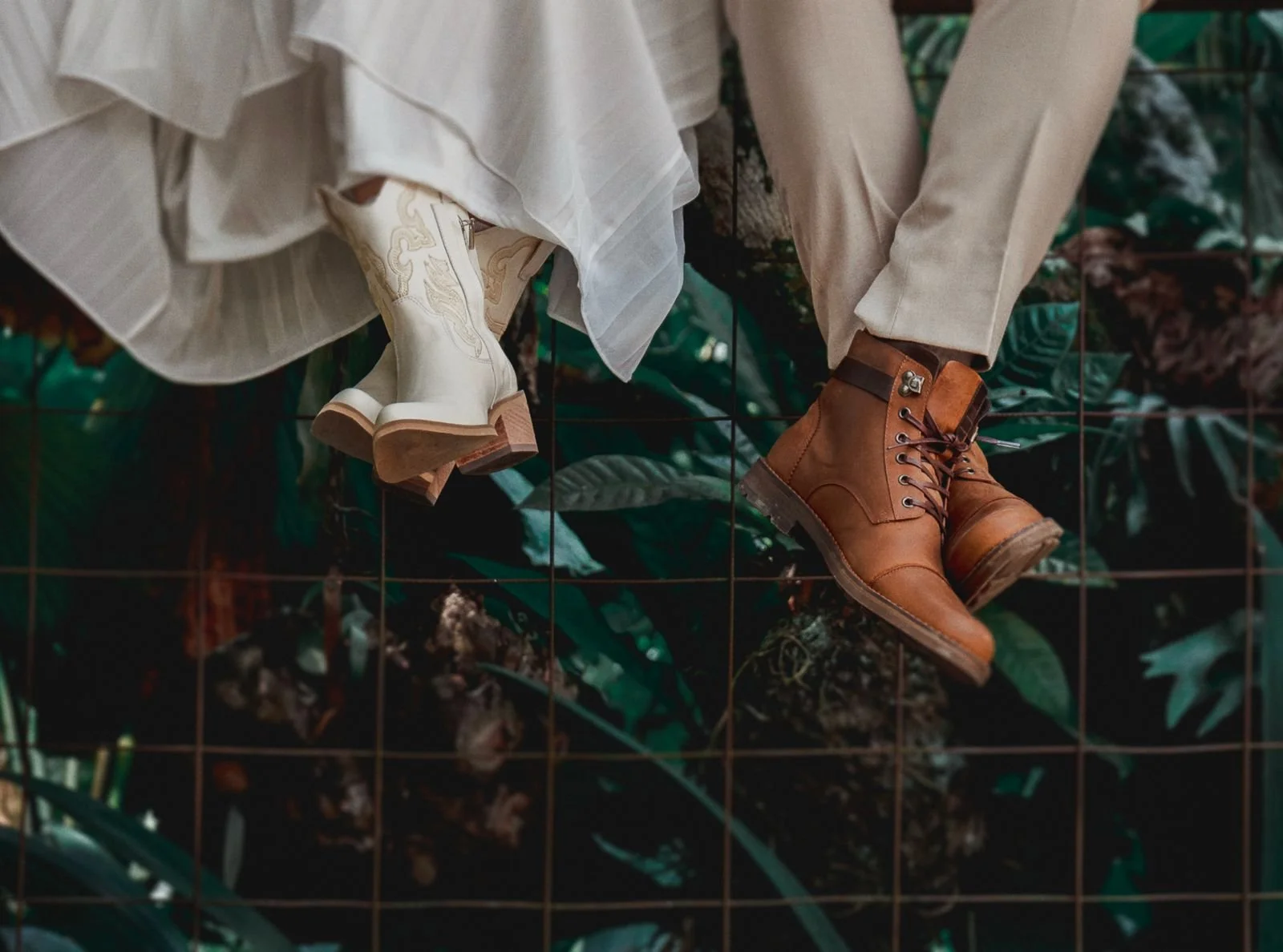 Zapatos de tacón y botas de cuero en una cerca de malla, con plantas verdes en el fondo.