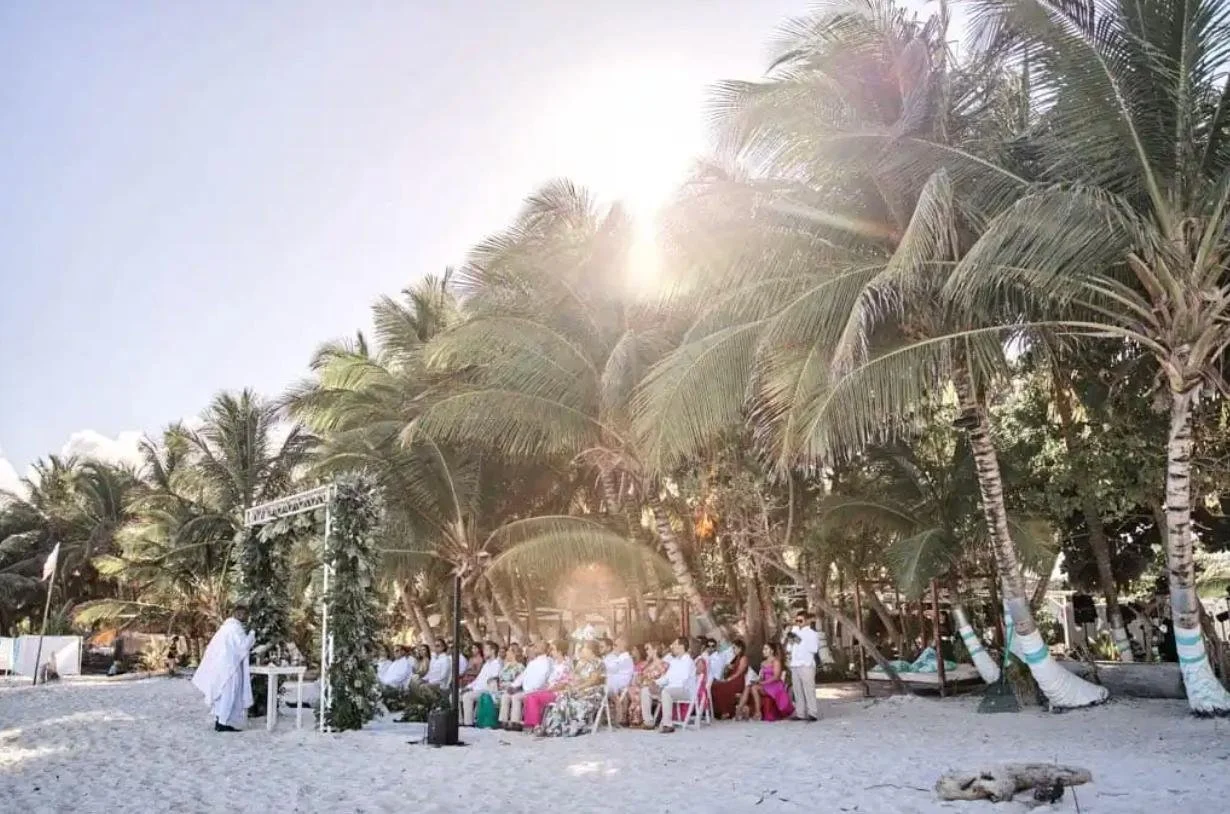 Ceremonia al aire libre en la playa con asistentes sentados bajo palmas, decoraciones y un oficiante frente a ellos, en un día soleado.