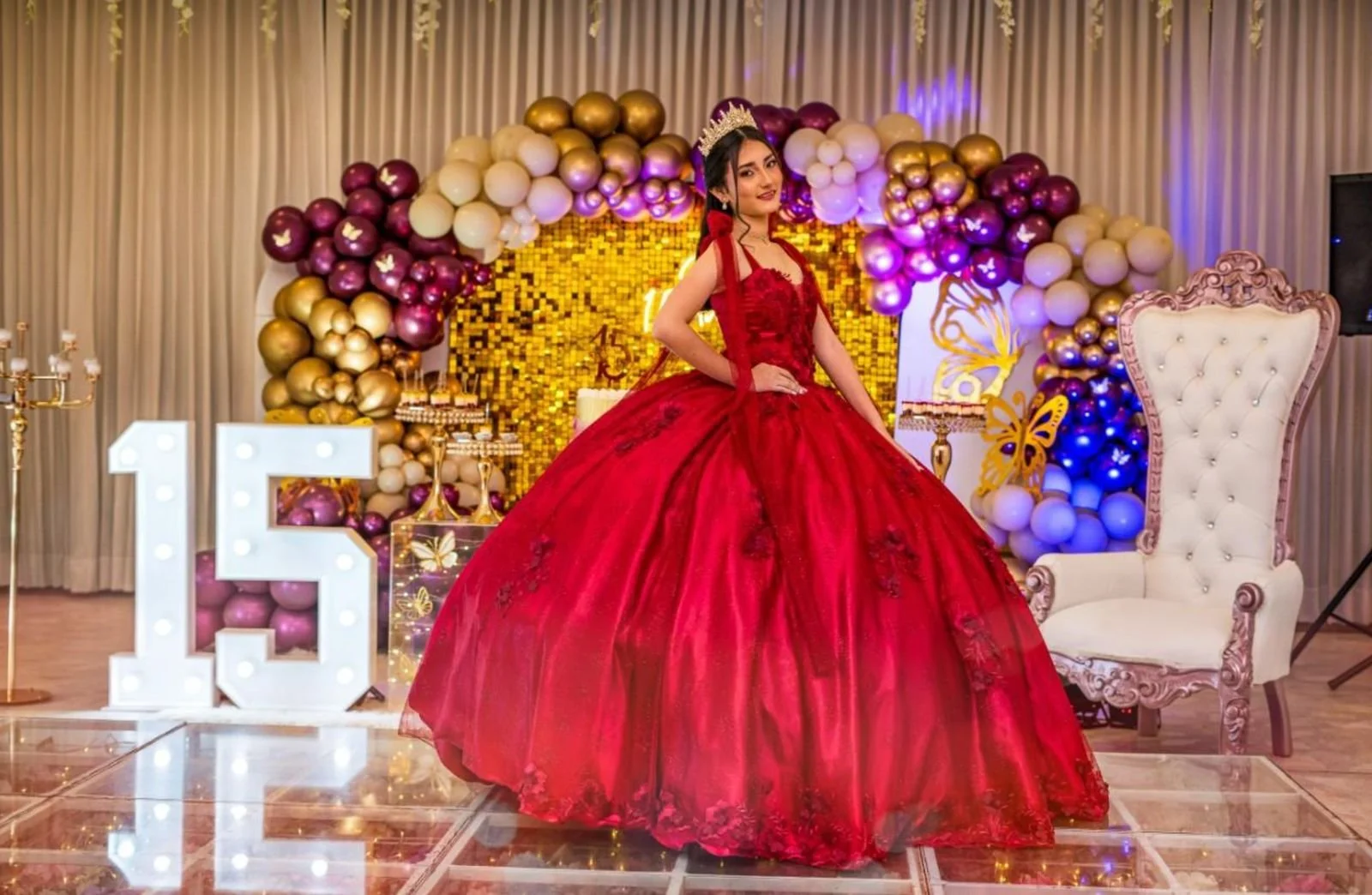 Joven en vestido rojo en fiesta con decoraciones doradas y blancas, globos y letras de luz en forma de número 15.