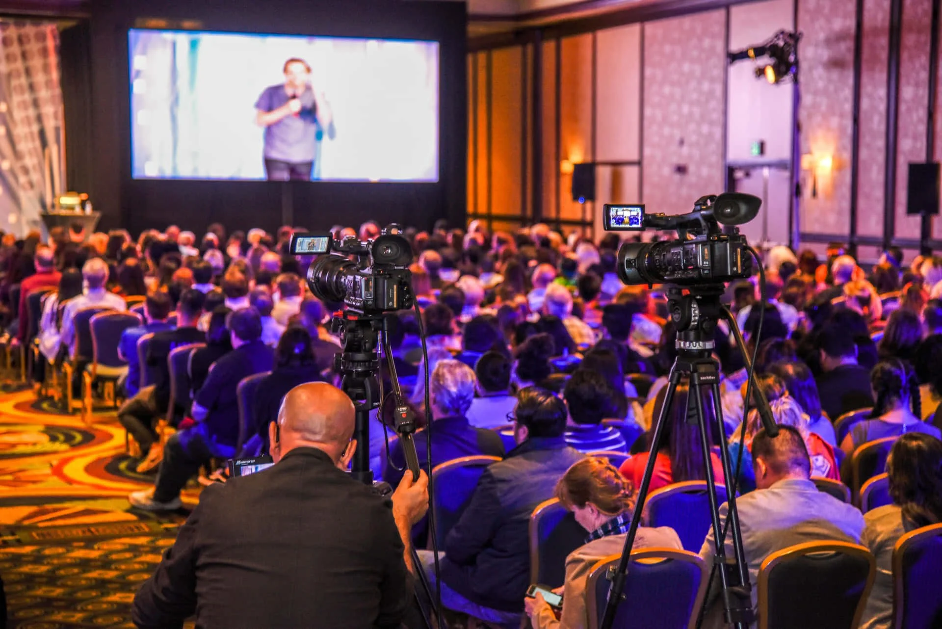Audiencia en un evento con cámaras de video enfocadas en el escenario y una pantalla grande con una persona hablando, en un lugar con paredes de ladrillo y iluminación cálida.