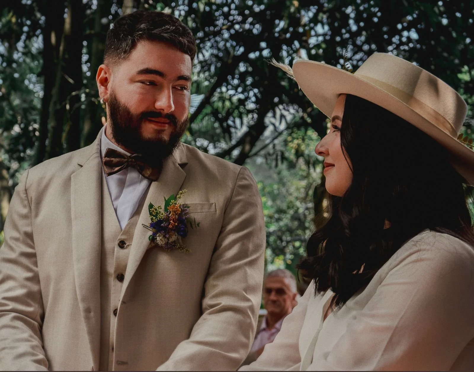 Una pareja en una boda al aire libre, con fondo de árboles y otras personas, en un momento emotivo, ambos con sonrisas y vestimenta formal.