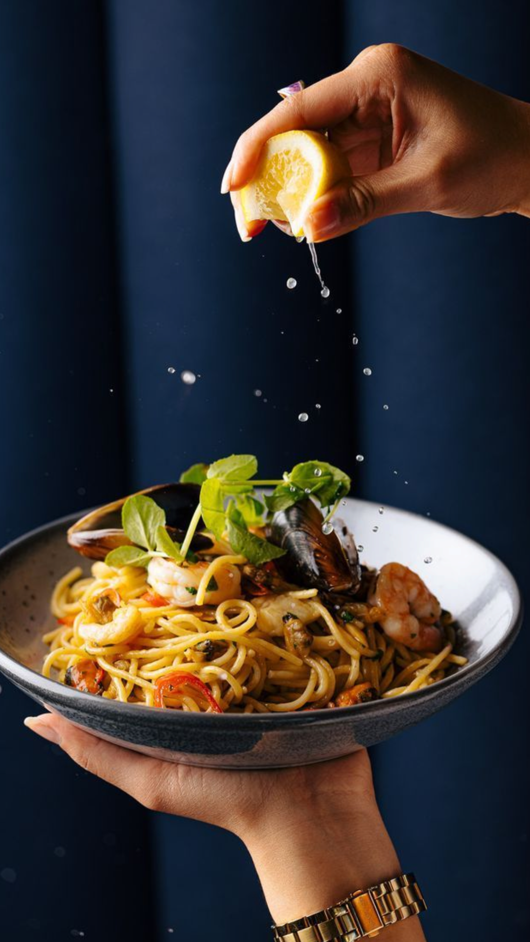 Mano rociando jugo de limón sobre un plato de pasta con mariscos y vegetales, fondo oscuro.