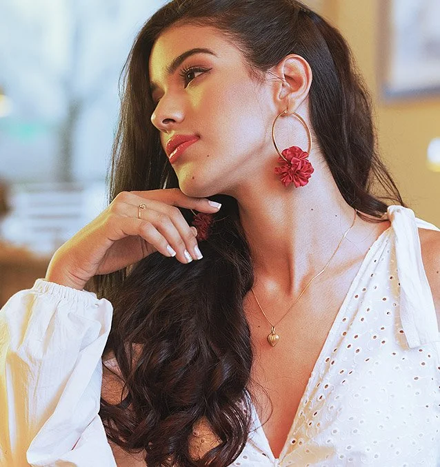 Mujer con cabello largo y oscuro, usando un vestido blanco, aretes grandes con flores rojas y collar con dije en forma de corazón, en un ambiente interior.