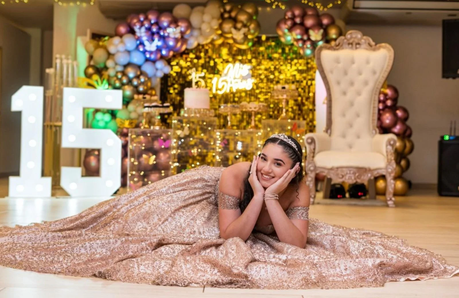 Una joven en un vestido elegante con corona, celebrando su quinceañera en un salón decorado con globos dorados y rosas, una gran letra 15 iluminada y una silla grande de estilo vintage.