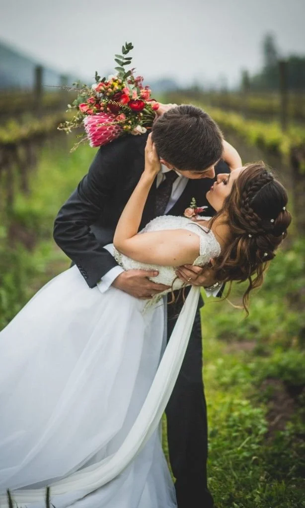Una pareja de novios en un campo, la pareja se está besando y la mujer está en brazos del hombre, ella lleva un vestido de novia y él usa un traje oscuro. La mujer sostiene un ramo de flores en su cabeza.