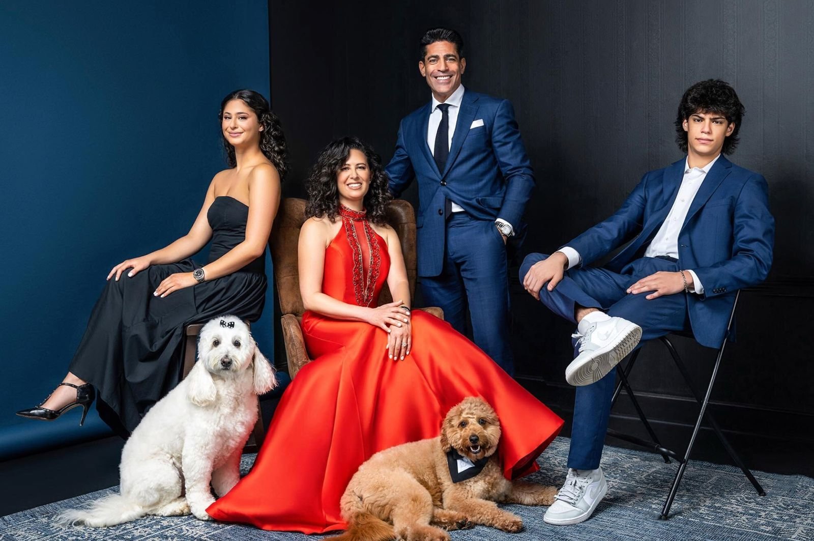 Grupo de cinco personas vestidas elegantemente con dos perros y un perro de peluche en un fondo oscuro.