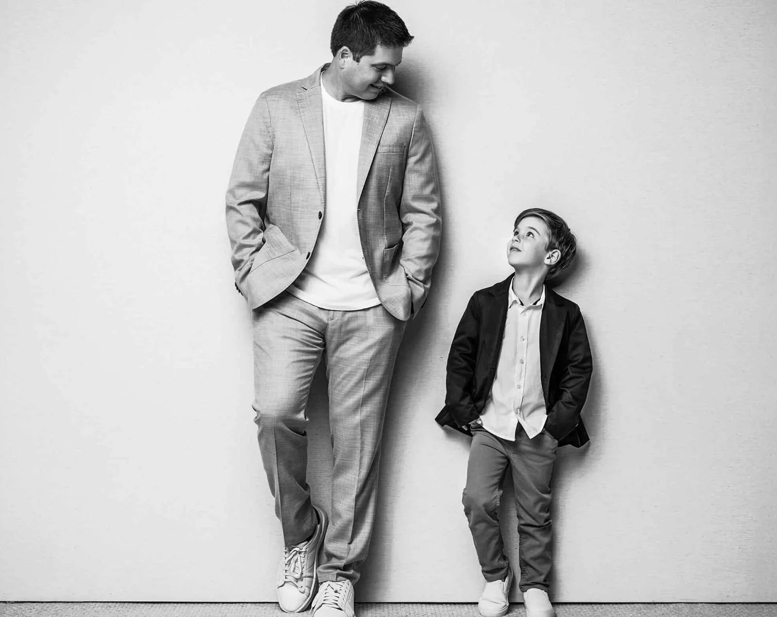 Hombre y niño de pie junto a una pared blanca, el hombre está sonriendo y mirando al niño, llevan ropa formal y están en tonos de gris