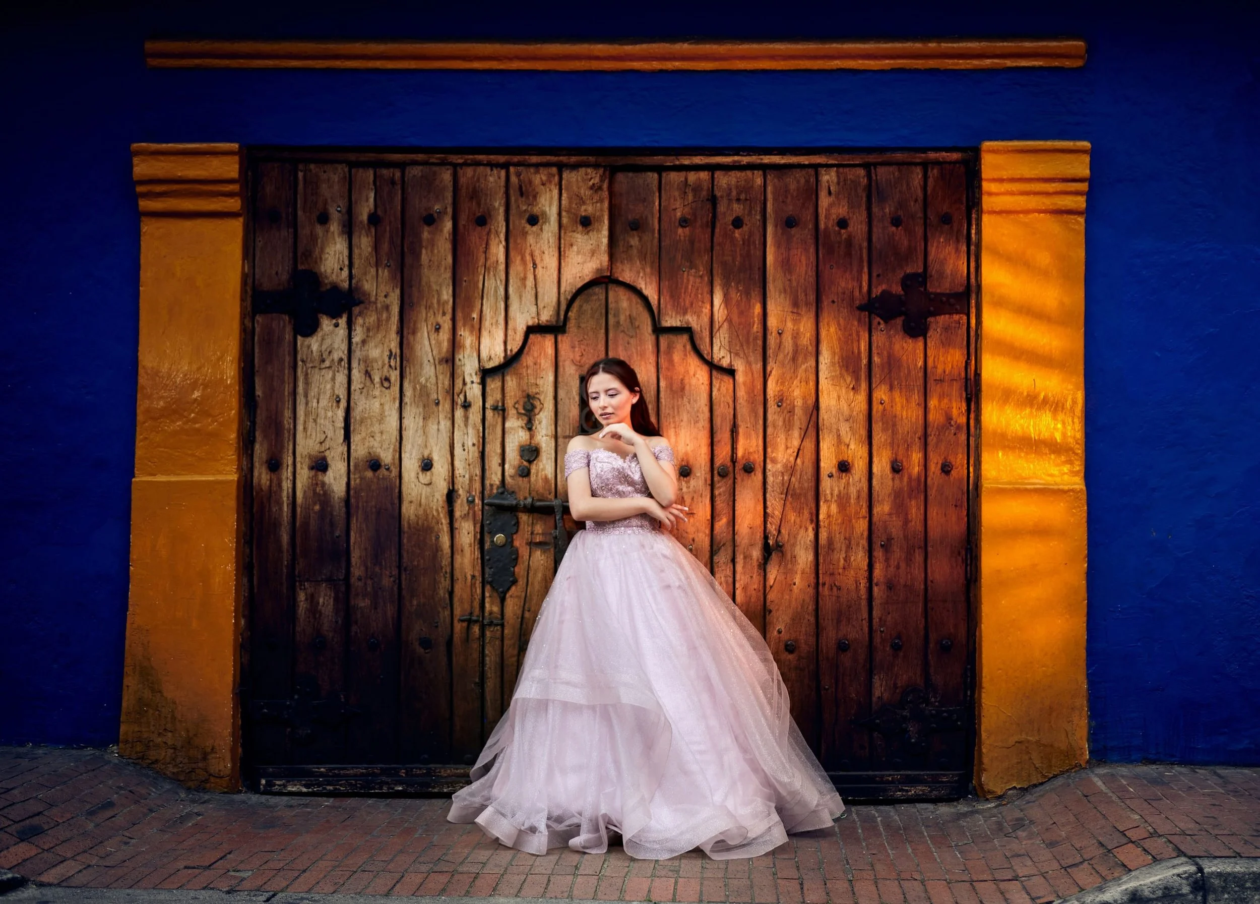 Mujer con vestido de quinceañera rosa, de pie frente a una puerta de madera grande y antigua, con paredes pintadas de azul y amarillo.