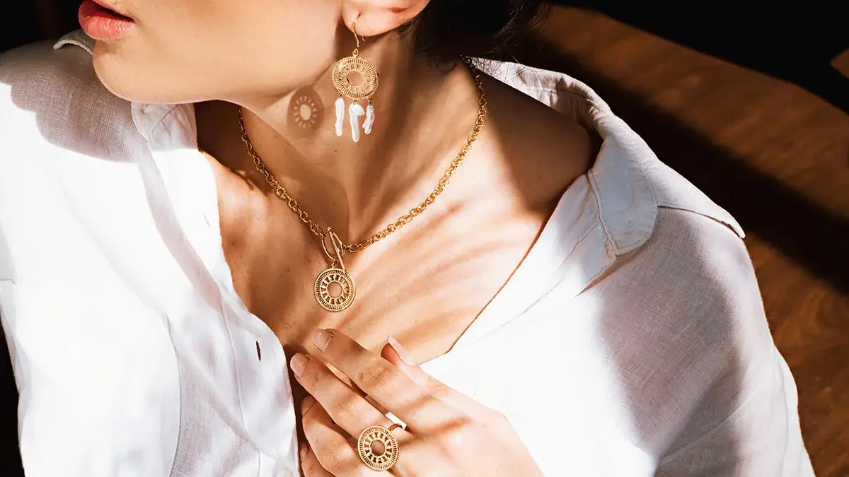 Mujer con pendientes, collar y anillo de joyería de color dorado, con blusa blanca, tocándose el pecho.