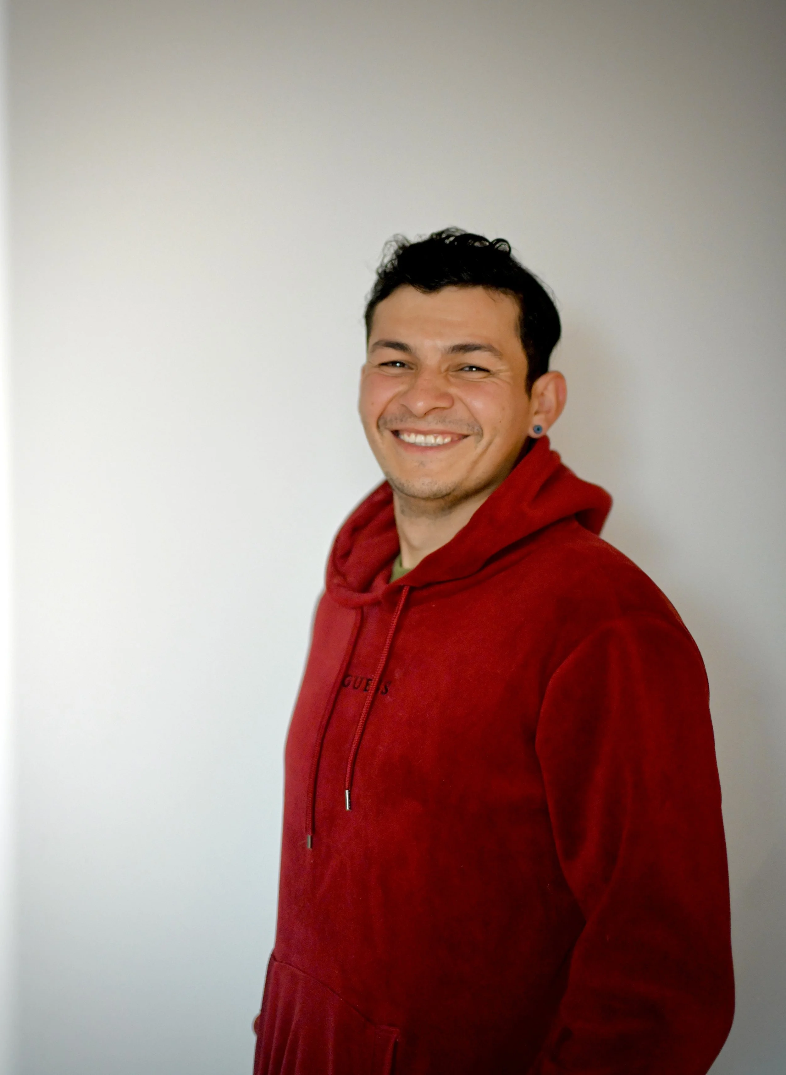 Hombre joven sonriendo, lleva sudadera roja, fondo gris claro.