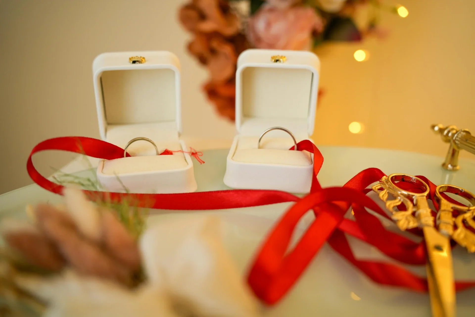 Dos anillos de bodas en cajas blancas decoradas con cinta roja, rodeados de accesorios dorados y lazos rojos, con decoraciones navideñas en el fondo.