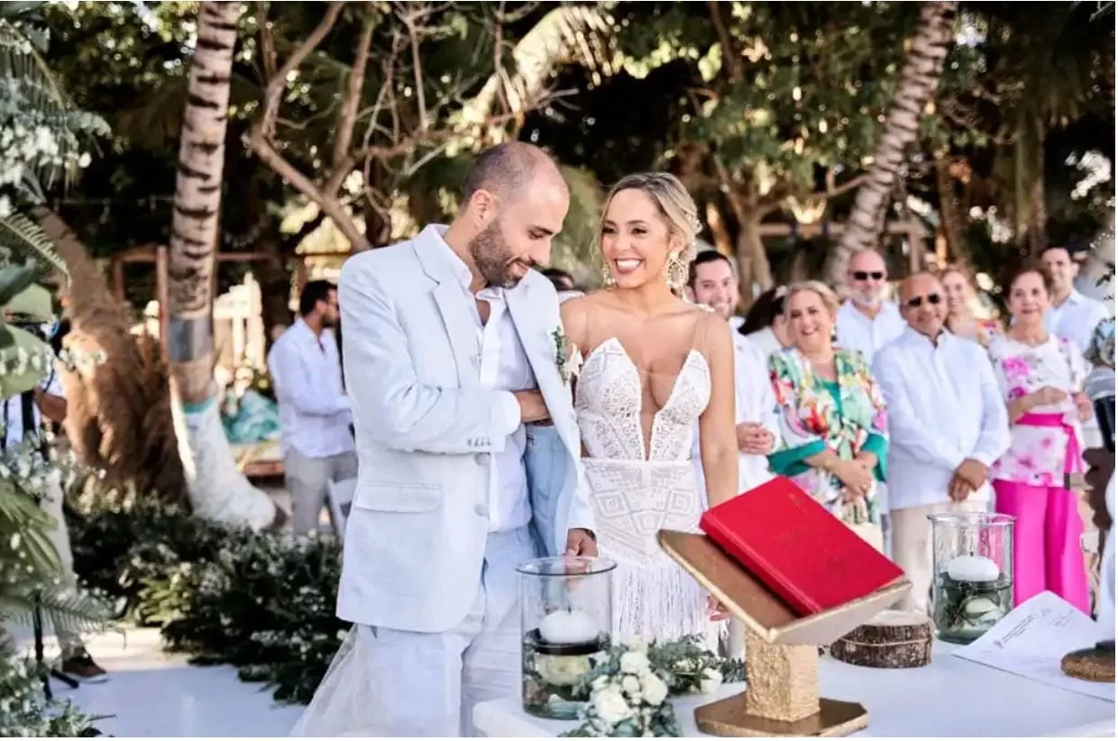 Pareja en una boda al aire libre con invitados de fondo, decoraciones florales y una mesa con libros y velas.