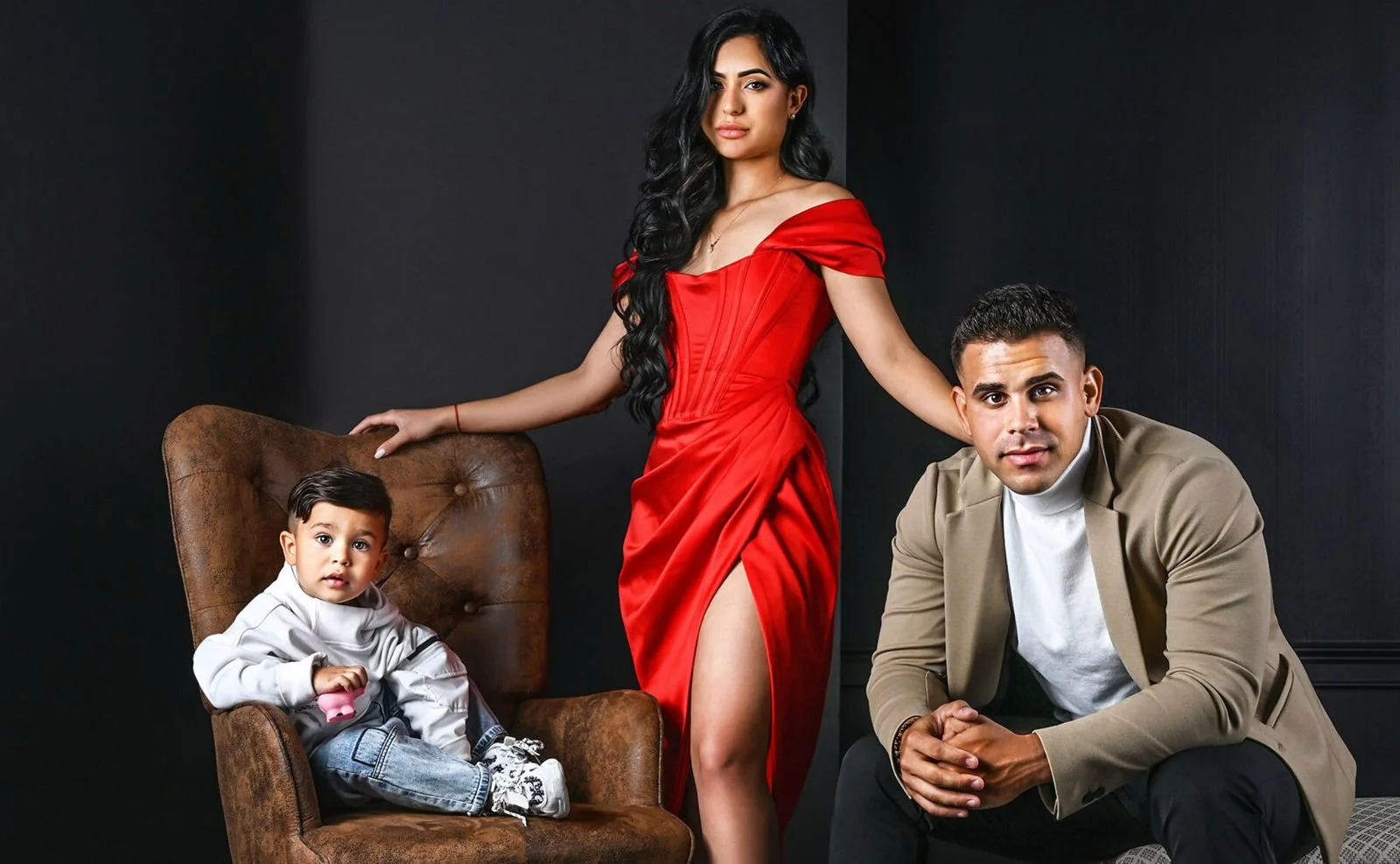 Una familia compuesta por una mujer, un hombre y un niño pequeño posando en un fondo oscuro. La mujer viste un vestido largo y rojo con una apertura en la pierna, el hombre lleva una chaqueta beige y camisa blanca, y el niño está sentado en una silla