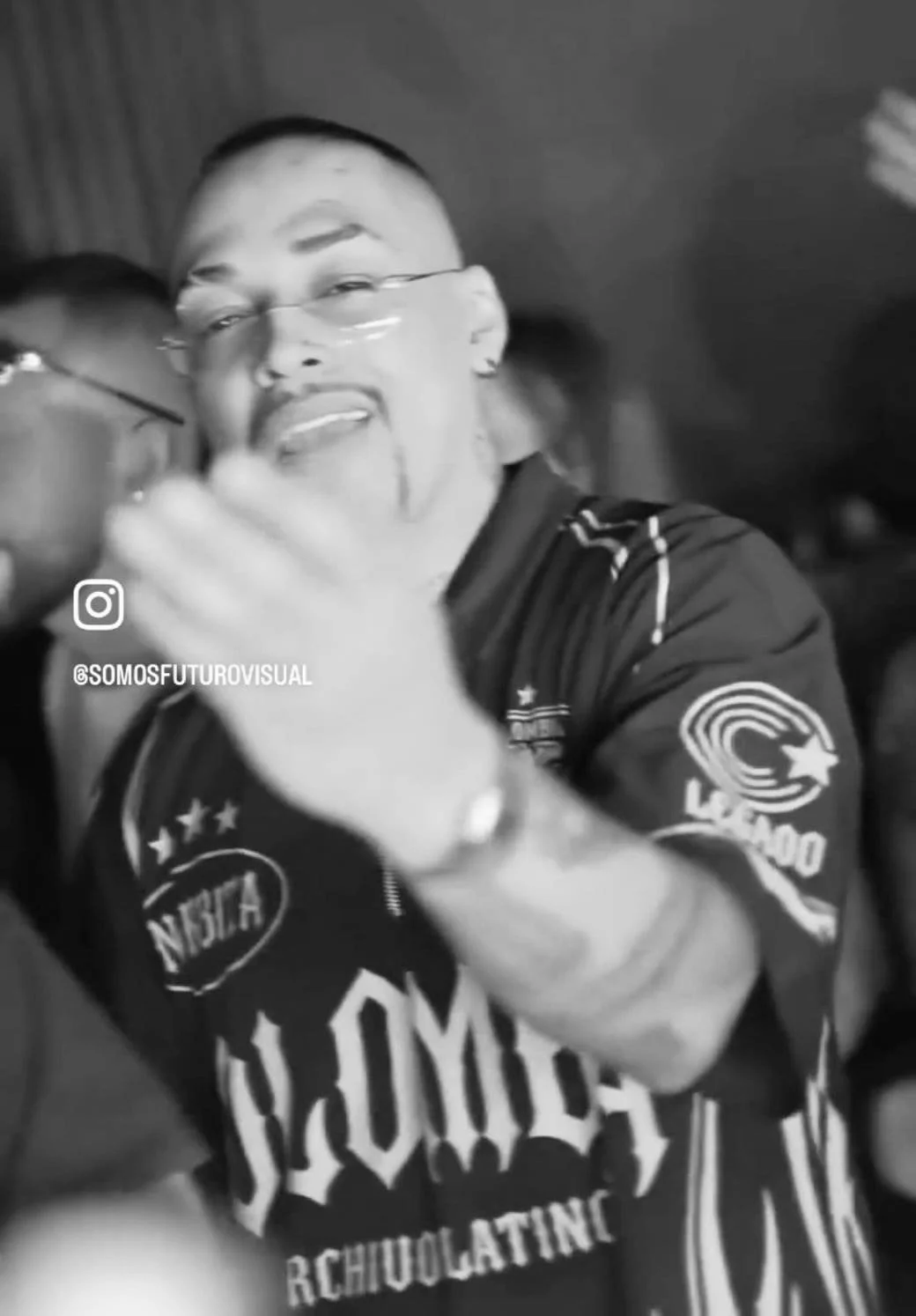 Persona sonriendo con gafas y camiseta negra con logos, en movimiento.