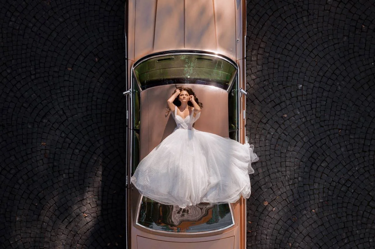 Mujer con vestido de boda blanca acostada sobre el techo de un coche de color cobre, en respuesta a una vista aérea, en una superficie de adoquines oscuros.