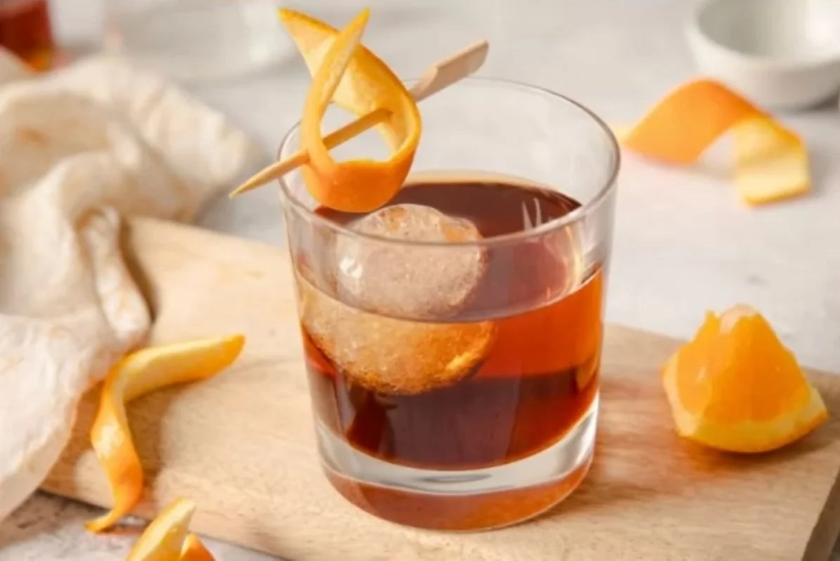 Un vaso con una bebida oscura, hielo y una rodaja de cáscara de naranja decorativa.