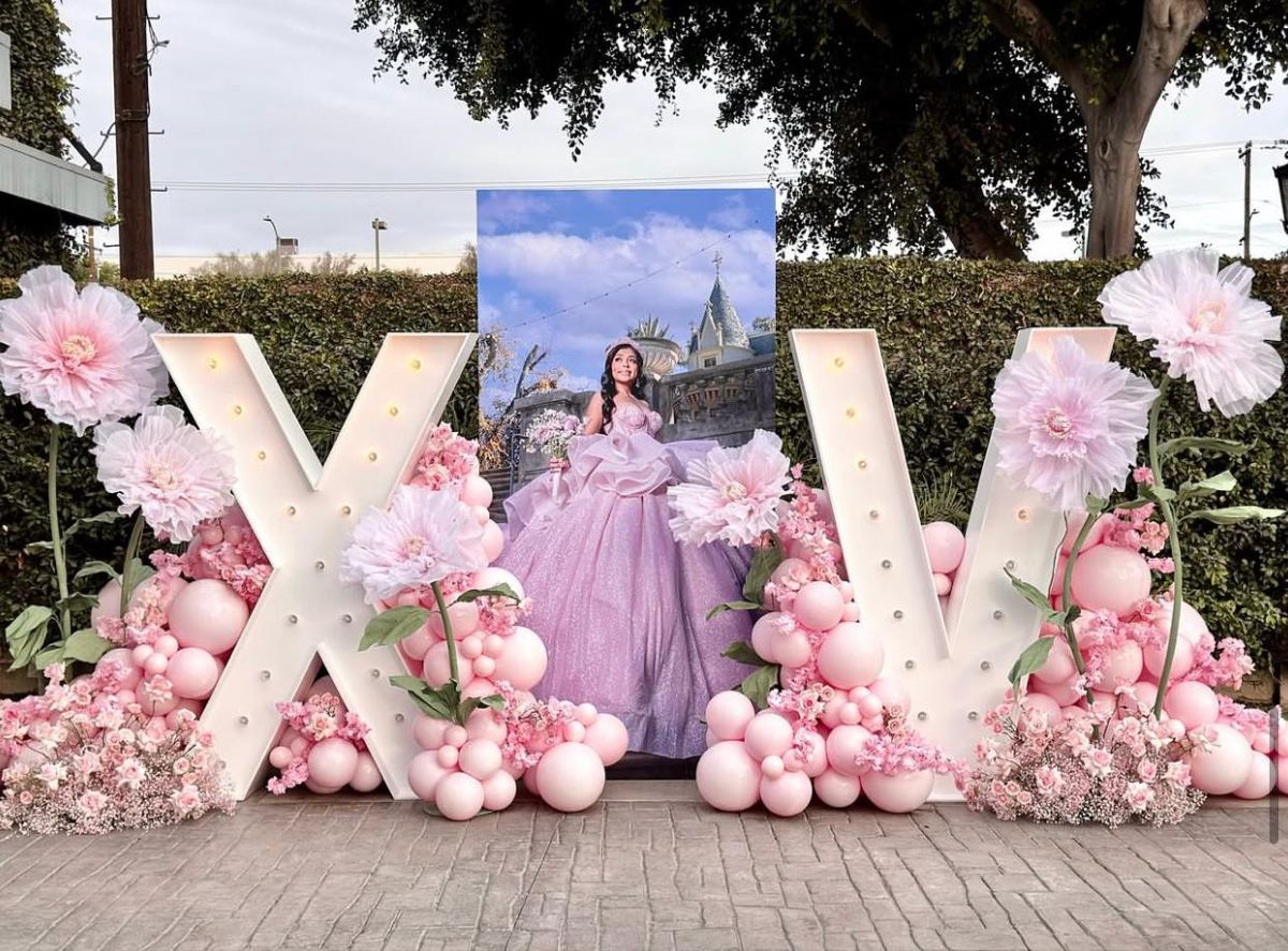 Decoración de letras grandes en forma de 'X' y 'V' con flores rosadas, globos rosados y una fotografía de una niña con vestido rosado en un evento al aire libre.