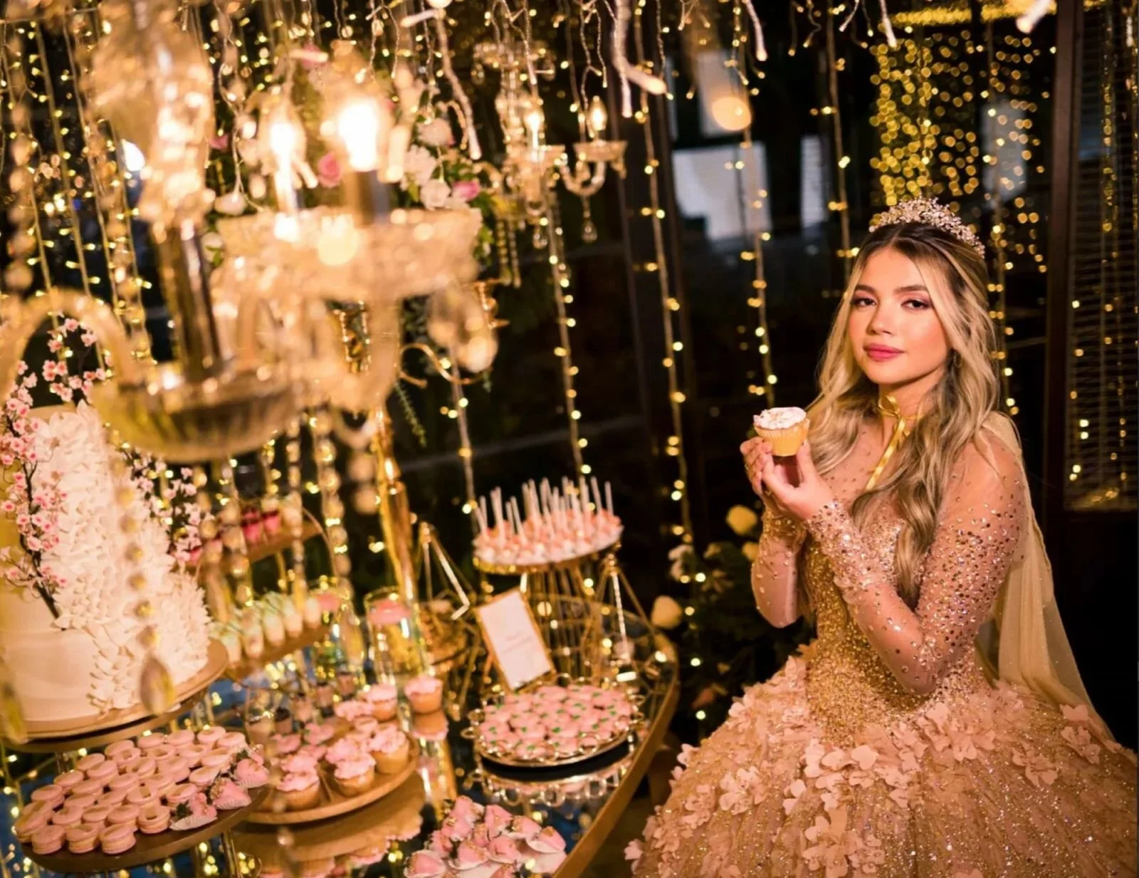 Mujer con vestido dorado y tiara en fiesta decorada con luces y arreglos florales, sosteniendo cupcake.