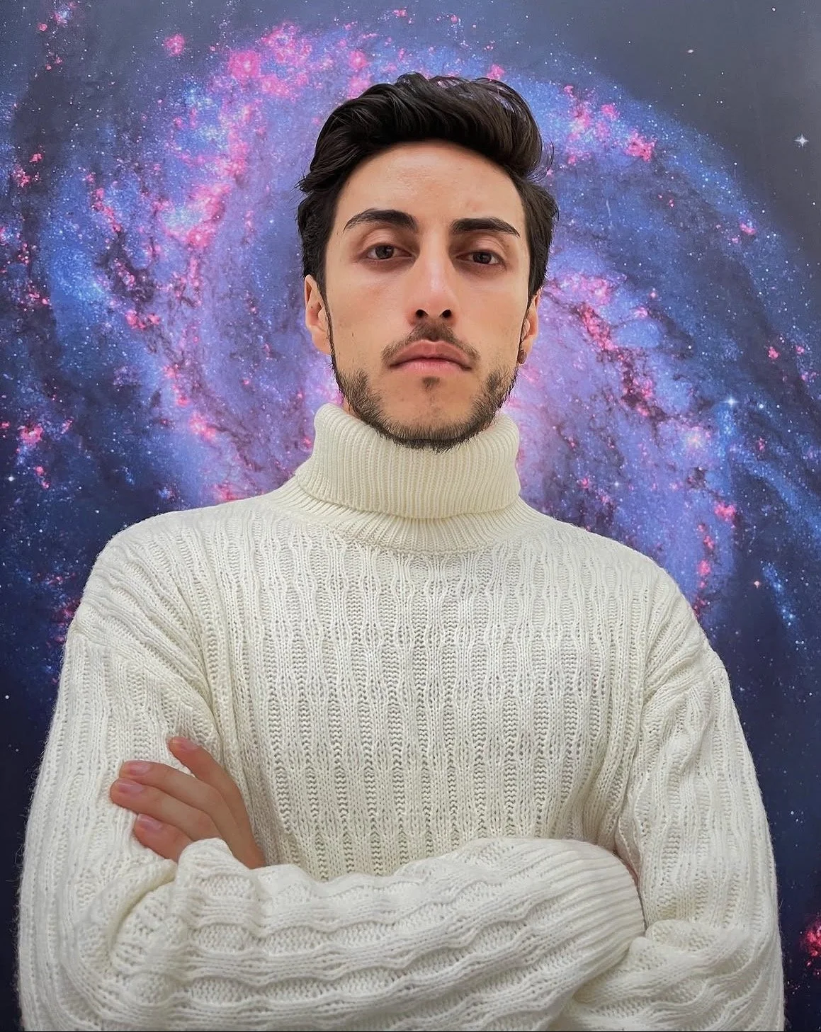 Joven con cabello oscuro, barba y cejas oscuras, usando suéter blanco de punto con cuello alto, frente a un fondo de galaxia con estrellas y nubes cósmicas.