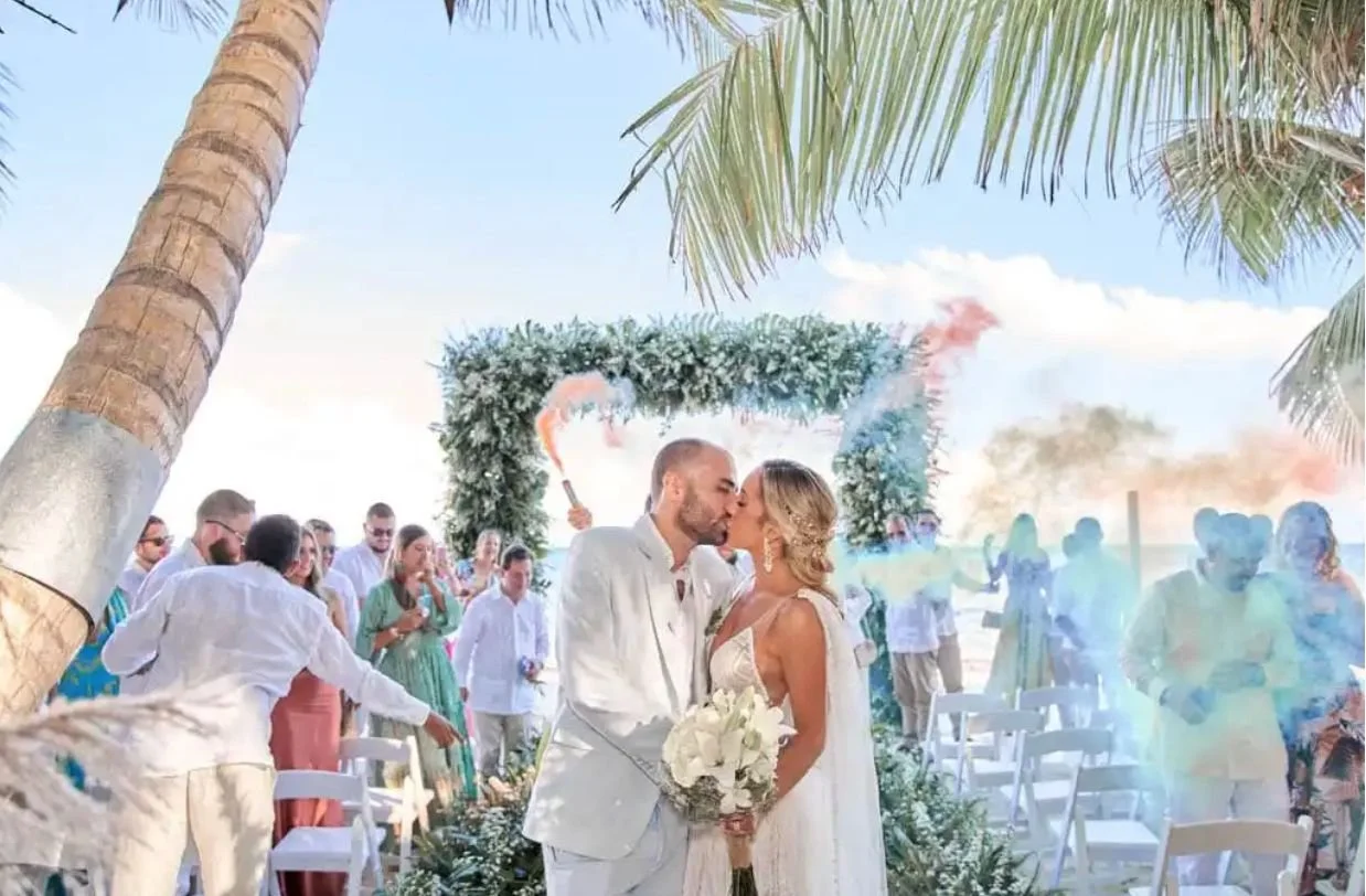 Pareja de recién casados besándose, en una ceremonia en la playa con invitados, decoraciones florales, palmeras y paisaje marítimo de fondo