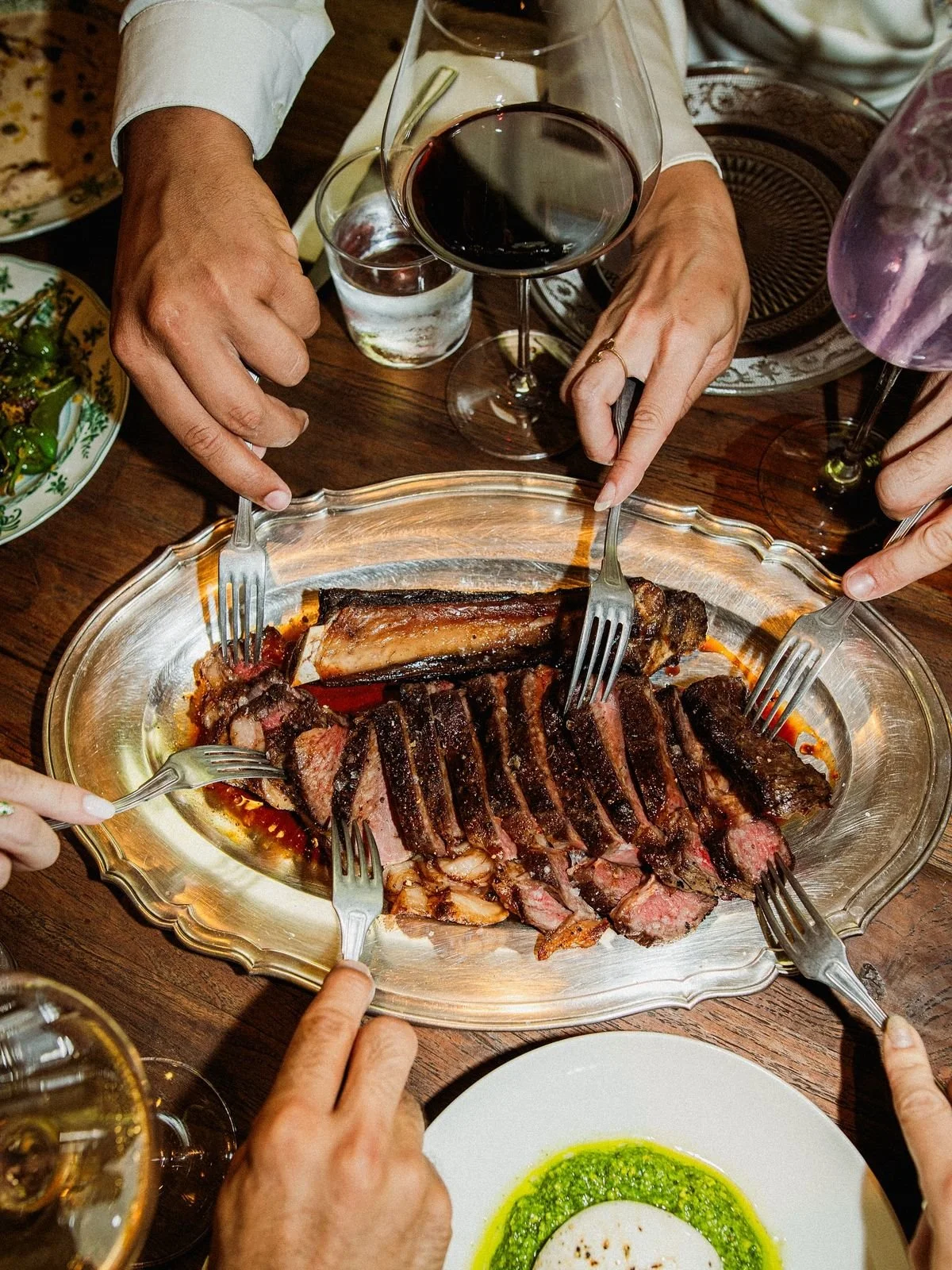 Varios tenedores y manos rodean un plato grande con carne asada en una mesa. También hay copas de vino y otros platos con comida.