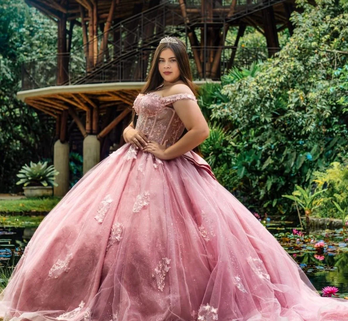 Mujer en vestido de ocasión formal rosado en un entorno natural con un árbol de gran tamaño y un estanque con flores acuáticas.
