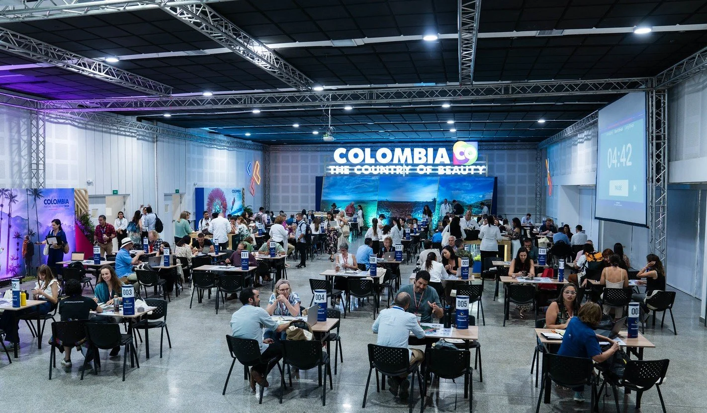 Evento en un salón grande con muchas personas sentadas en mesas, algunas de ellas conversando, otras usando laptops. En el fondo hay una pantalla grande con el texto 'Colombia, el país de la belleza' y diversas decoraciones relacionadas con Colombia,